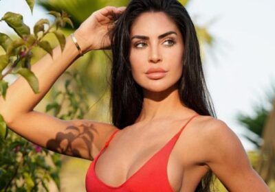 Woman with long dark hair in a red bikini top posing outdoors among tropical plants in sunlight.